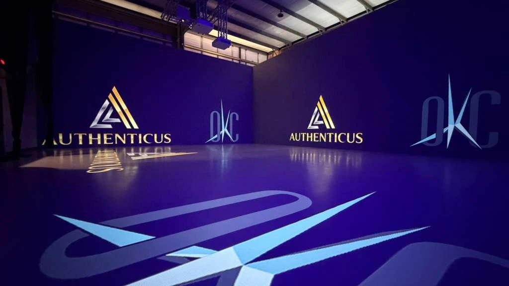 Immersive projection room with Authenticus and DKC visuals mapped across two walls and the floor, with ceiling-mounted projectors