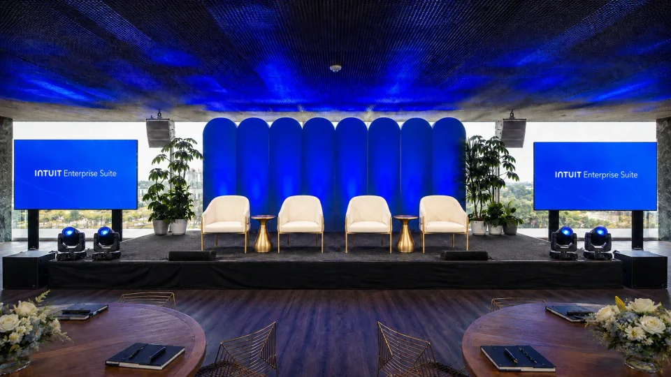 Retouched frontal view of the Intuit stage at POST Houston with four armchairs, branded LED displays, active lights and white hanging speakers